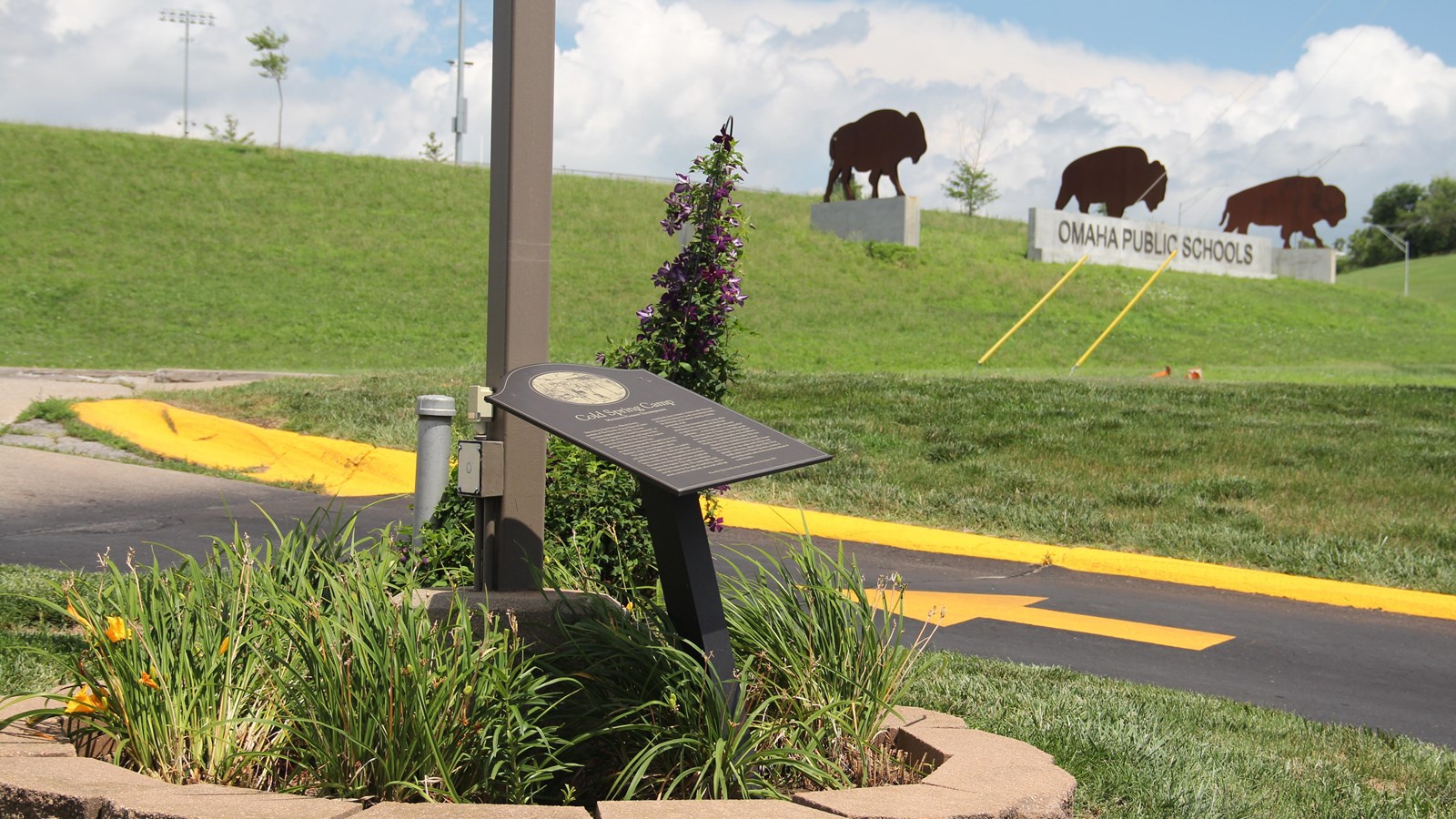 Historical marker in a garden with lilies, bison silhouettes, and Omaha Public Schools sign nearby.
