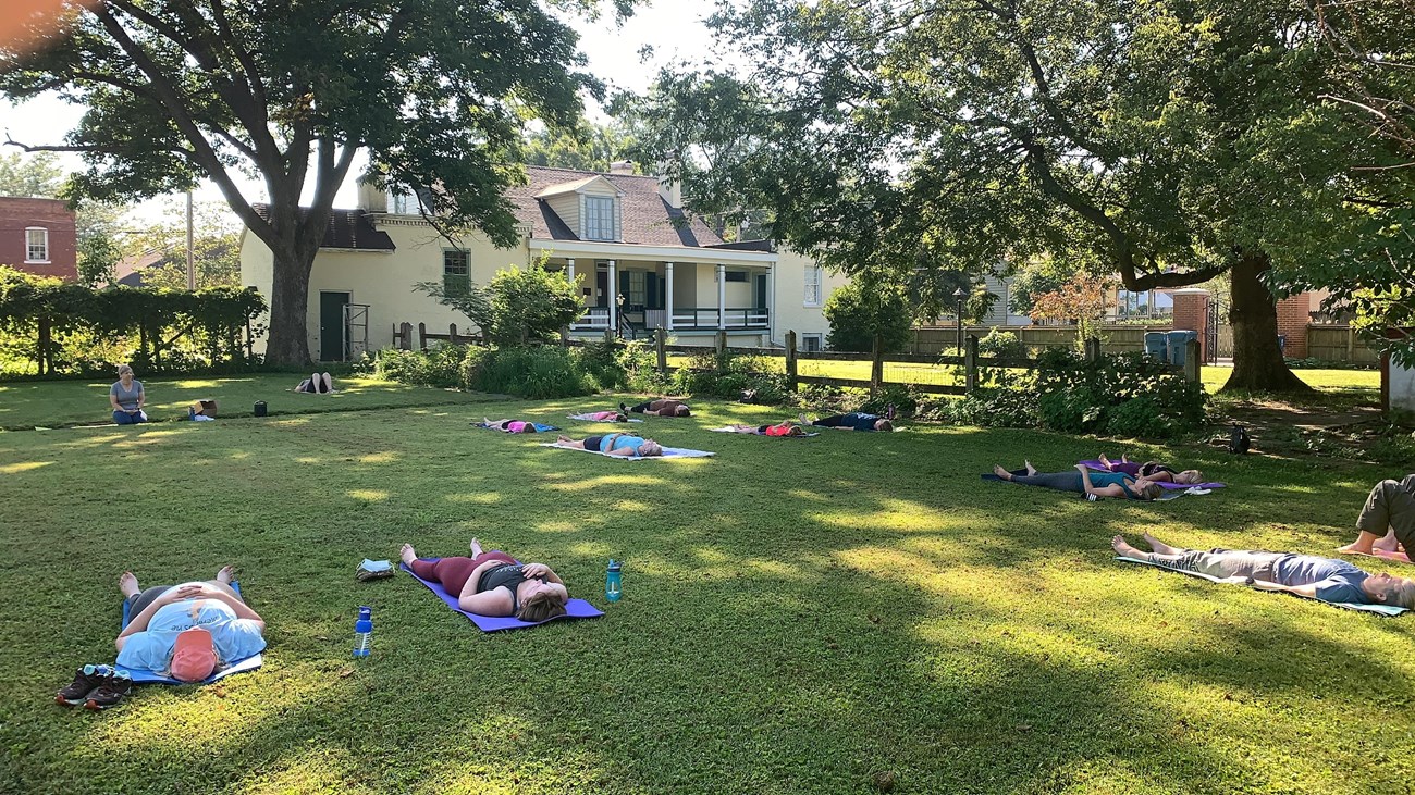A group of people laying on yoga mats in a large yard in behind a yellow historic house