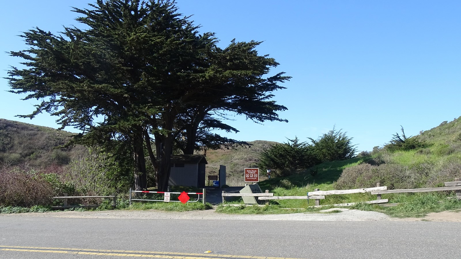A dirt trail passes by a gate, refuse containers, a trailhead sign, trees, and vault toilets.