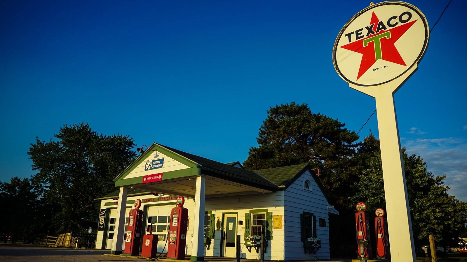 A small white sided building with a green roof. Out front are two red old-fashioned gas pumps.