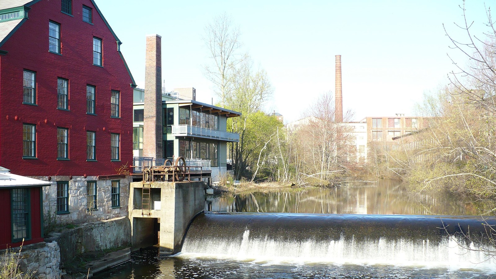 Red building along the banks of Mumford River with dam visible in center of shot