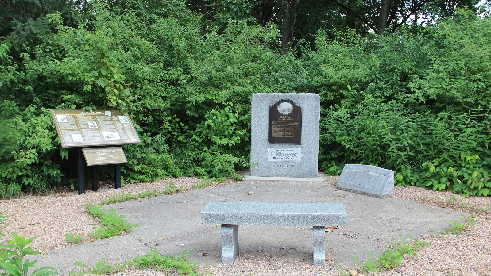 Square plaza with bench, granite monument, exhibits, and lush greenery at Cutler’s Park.
