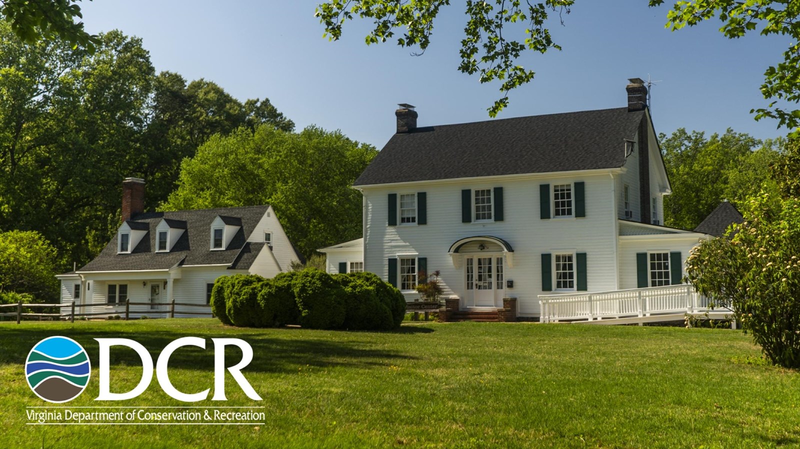 A white two-story house and a adjacent building sits on a green lawn, surrounded by trees.