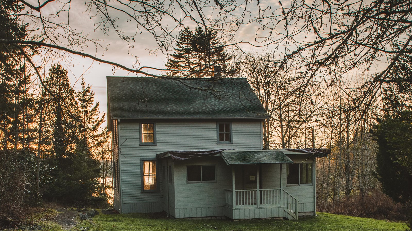 Color photograph of a frame house with porches in a green field at sunset