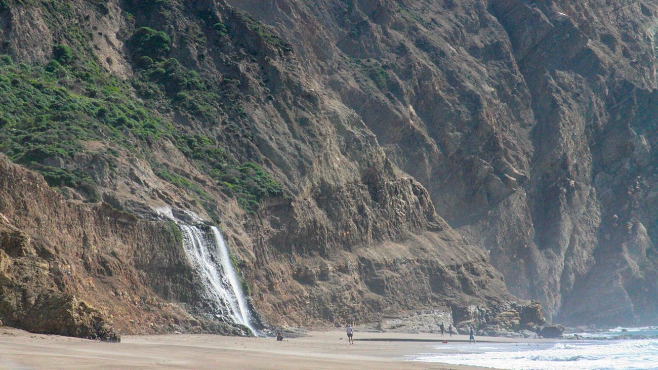 White water cascading over grass-topped coastal bluffs to a sandy ocean beach with four people below