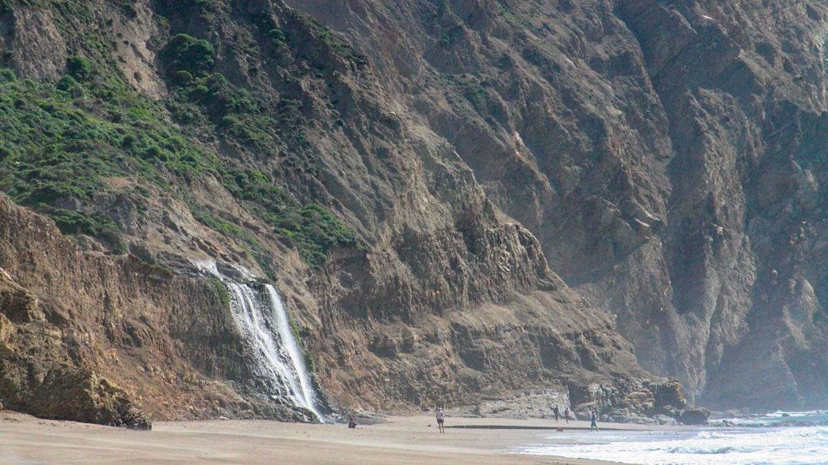 White water cascading over grass-topped coastal bluffs to a sandy ocean beach with four people below