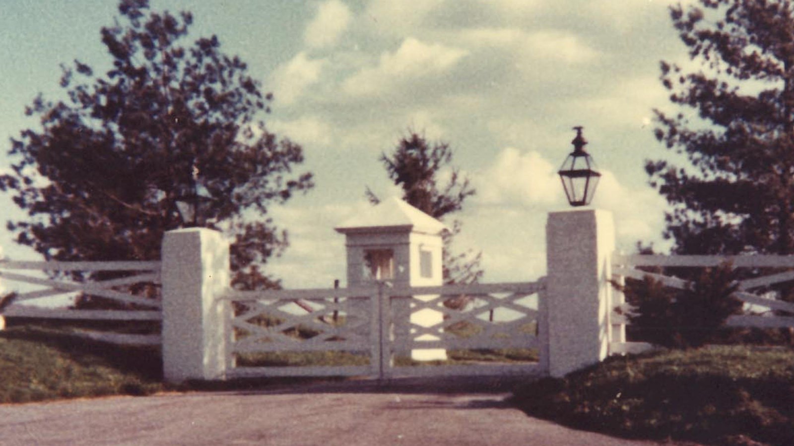 A white, wood gate is flanked by white brick columns. Behind the gate is a small, square, white hut.