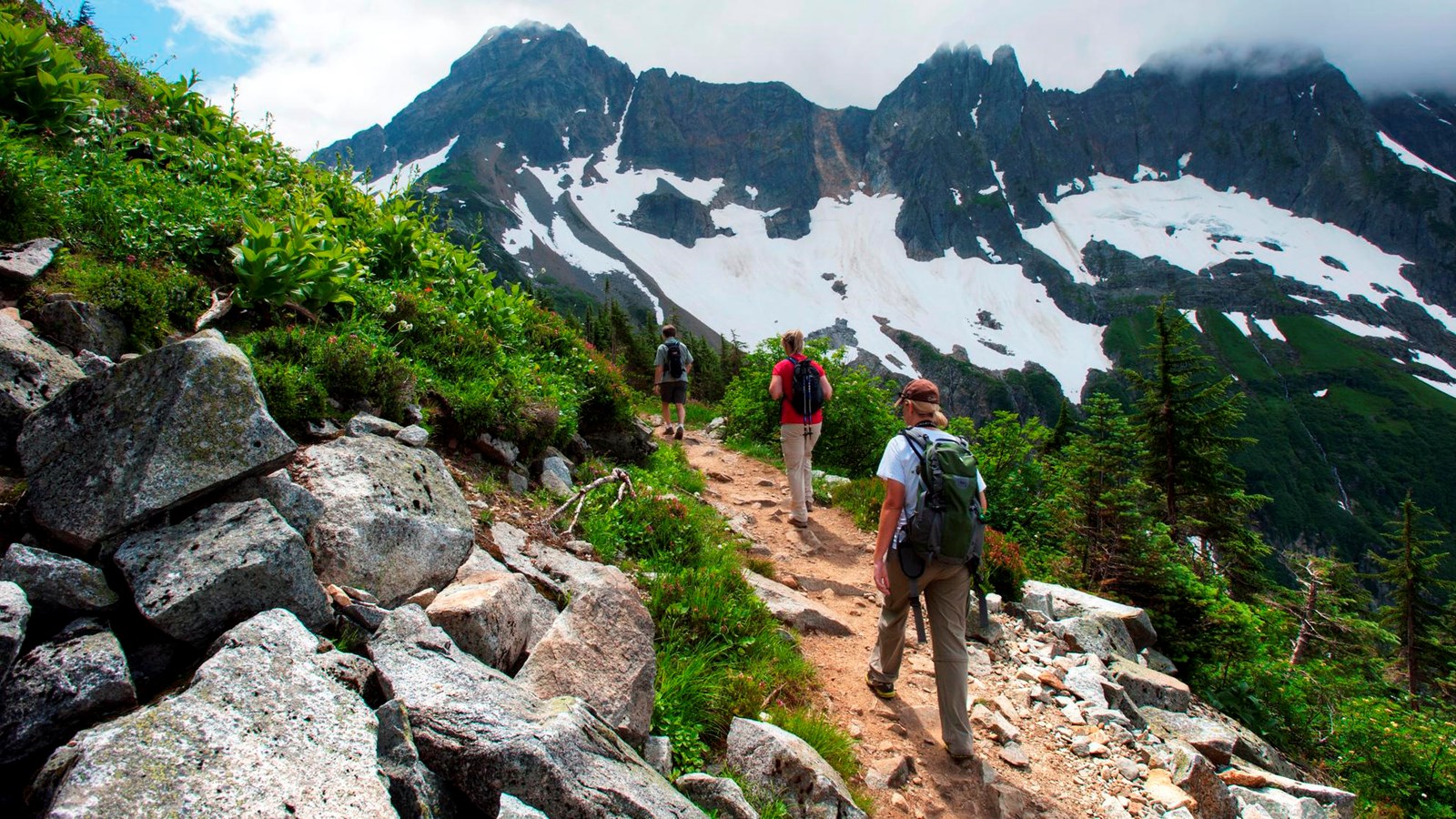 Three hikers travel a dirt path on the side of a steep grassy slope, with mountains in the distance.