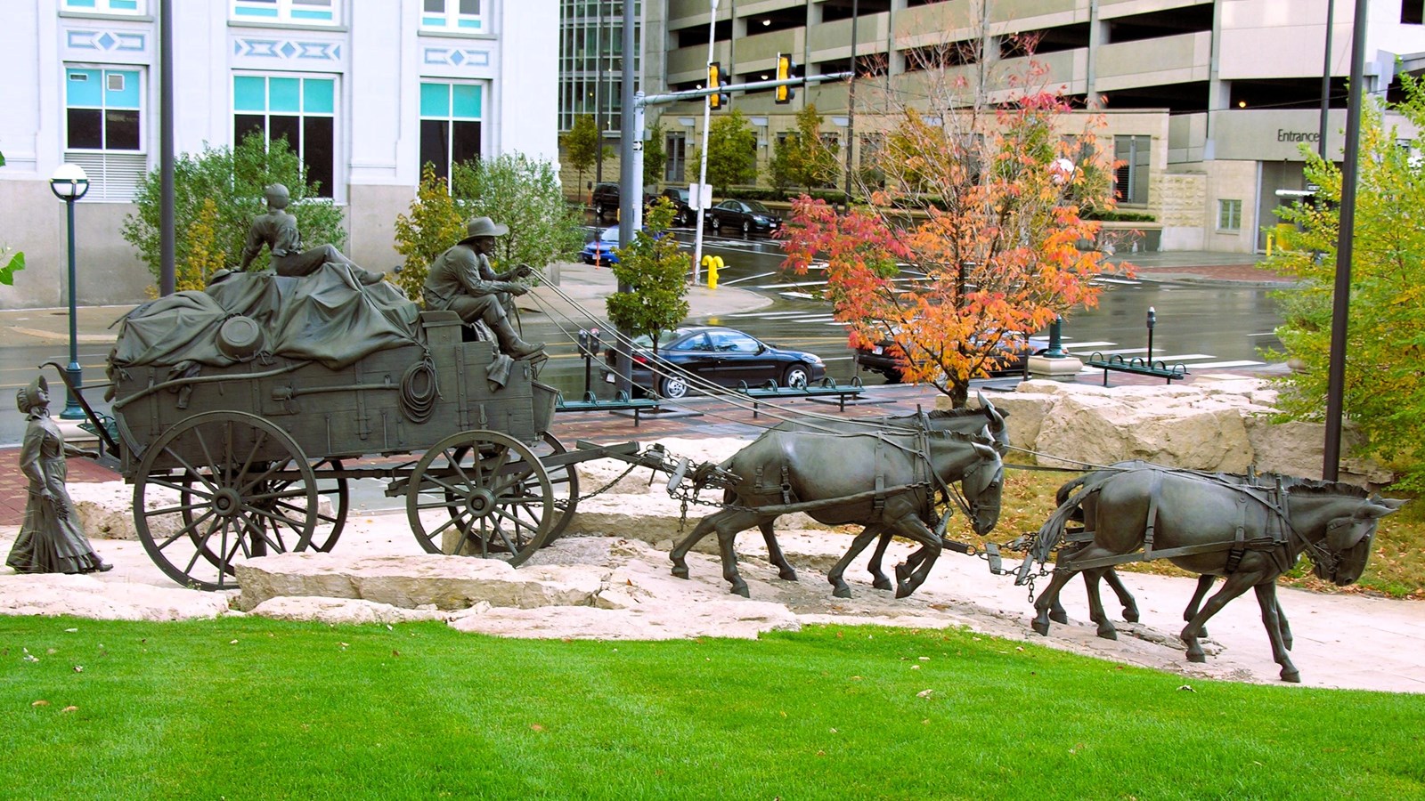 Larger-than-life mule-drawn wagon sculpture on grass, Omaha city street and buildings behind.