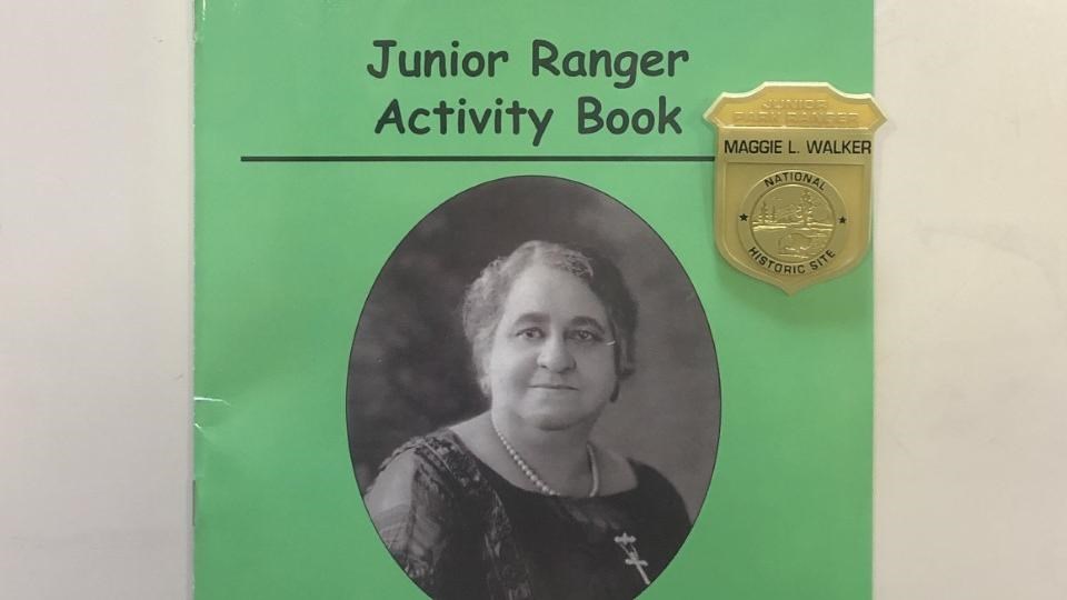 A Green booklet with a picture of Maggie L. Walker on front, a plastic badge sitting on the booklet