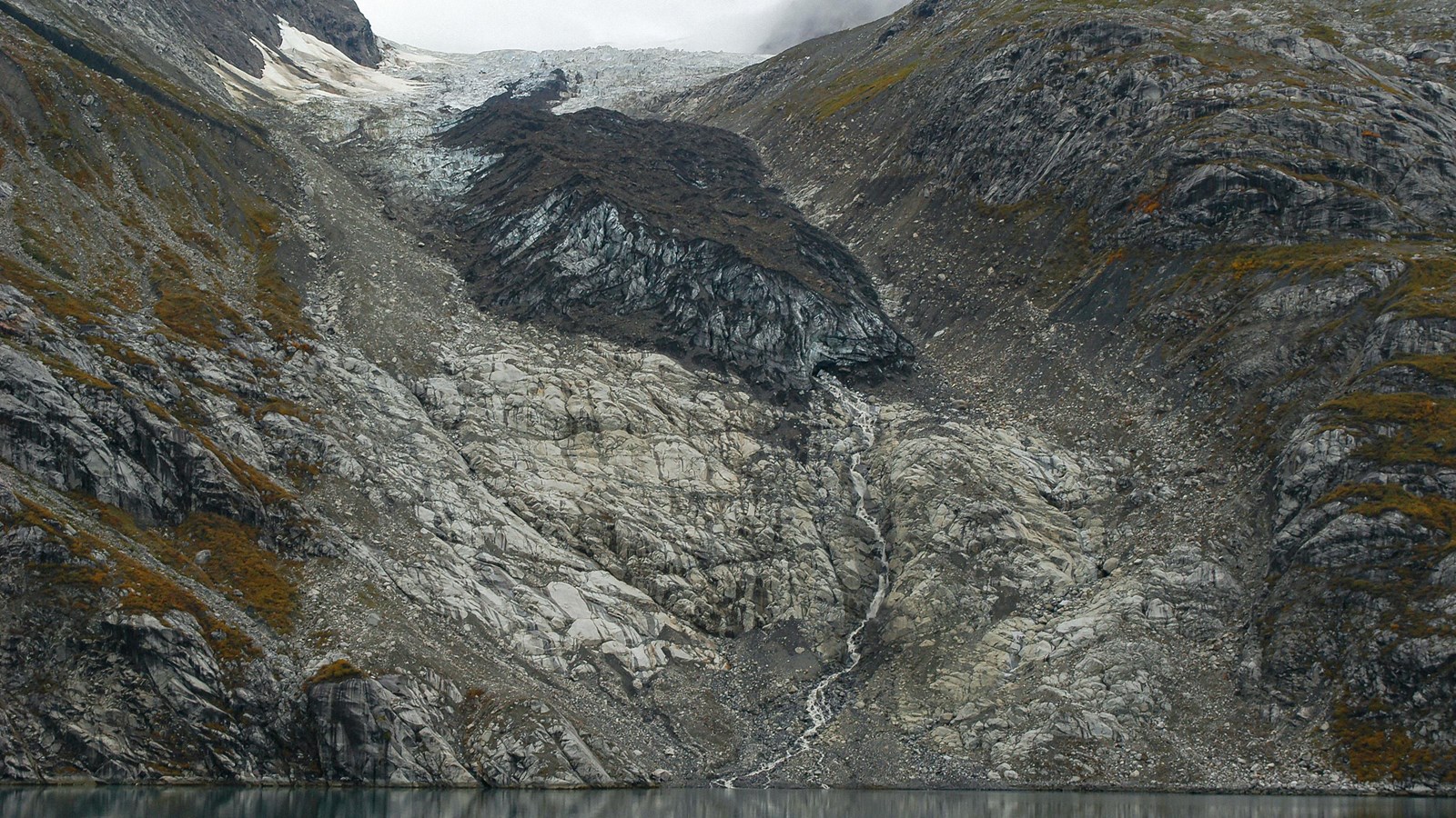 A glacier sits on a rocky hillside with a waterfall coming from its base.