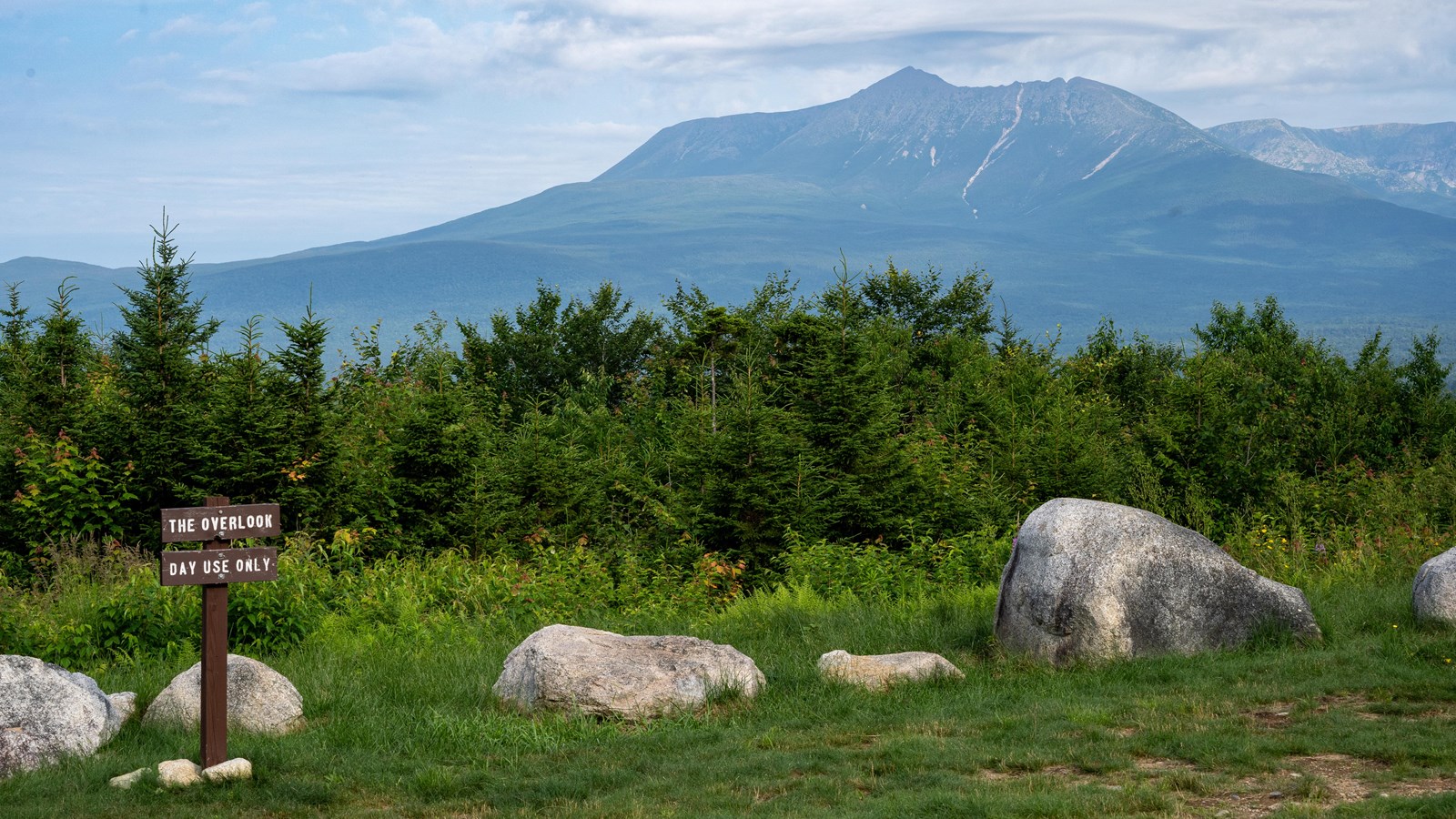 A green mountain peak rises above the treetops. In the foreground are boulders and grass.