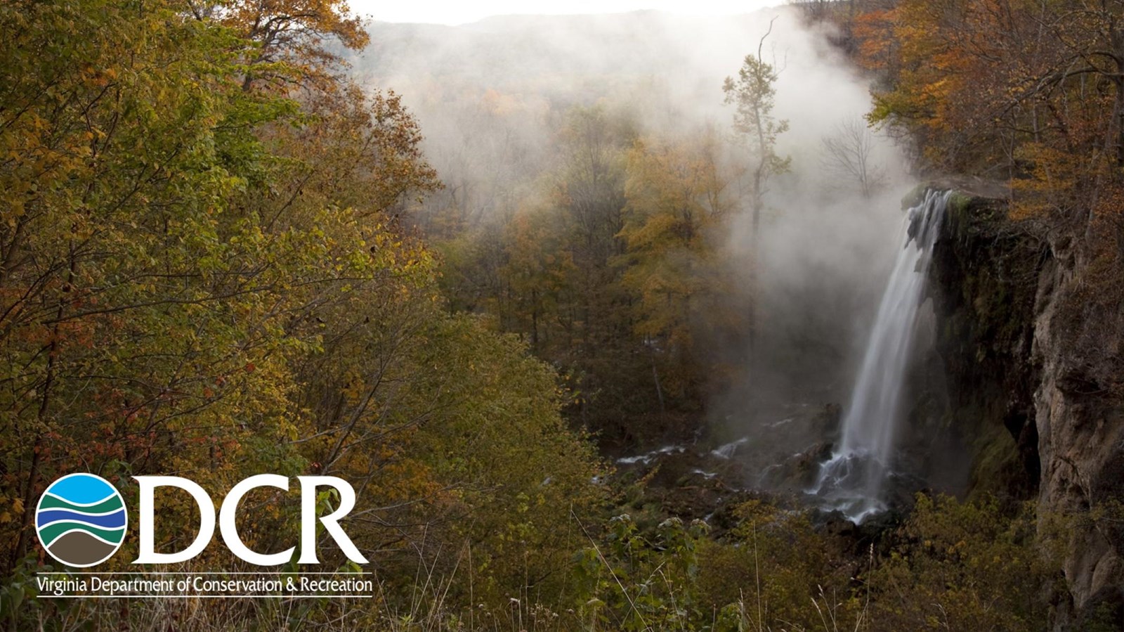 A waterfall cascades down a rocky cliff surrounded by autumn foliage, with mist rising into the air.