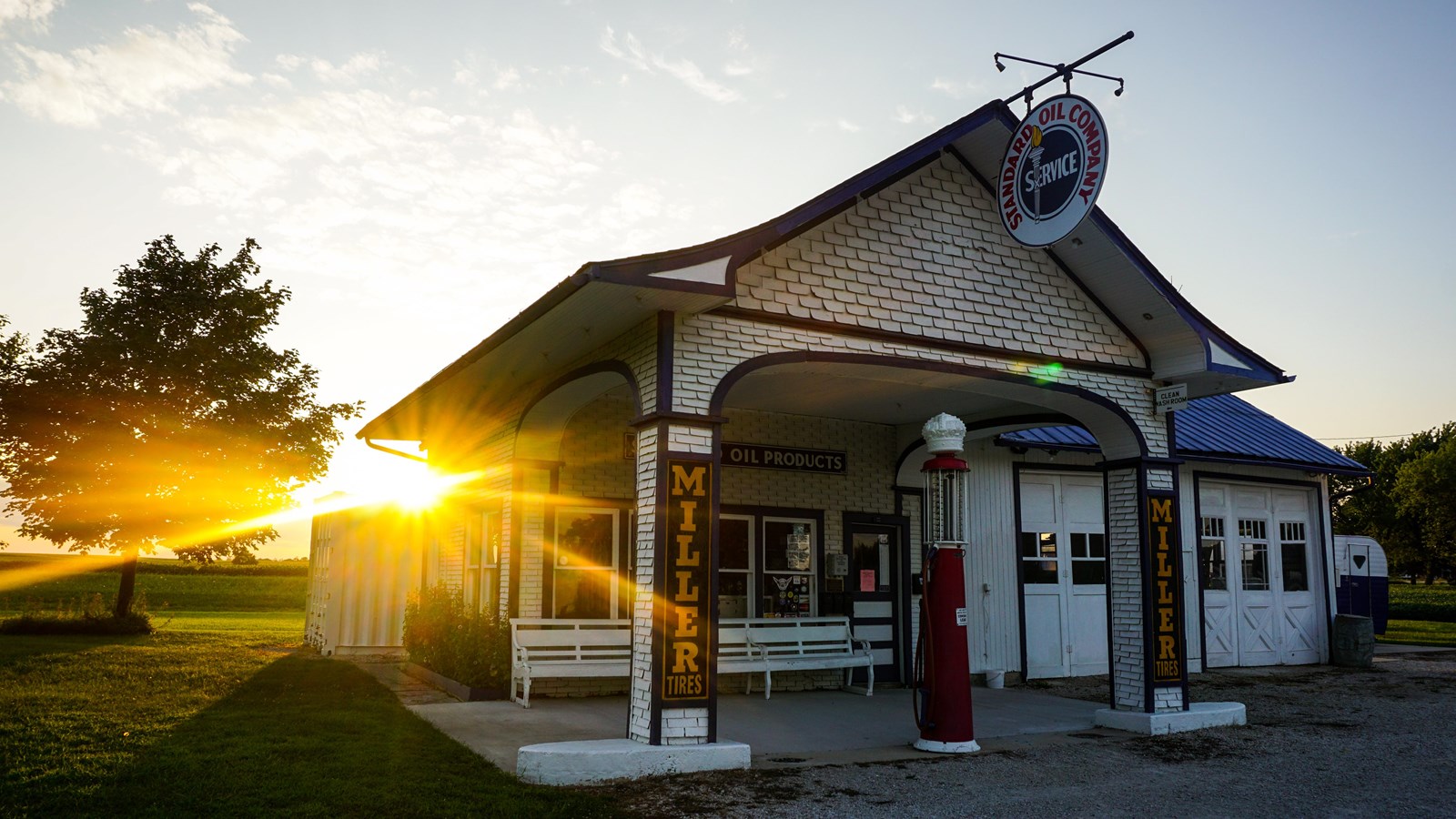 A white brick building with a grey roof and old-fashioned gas pump stands with rays of sun behind.