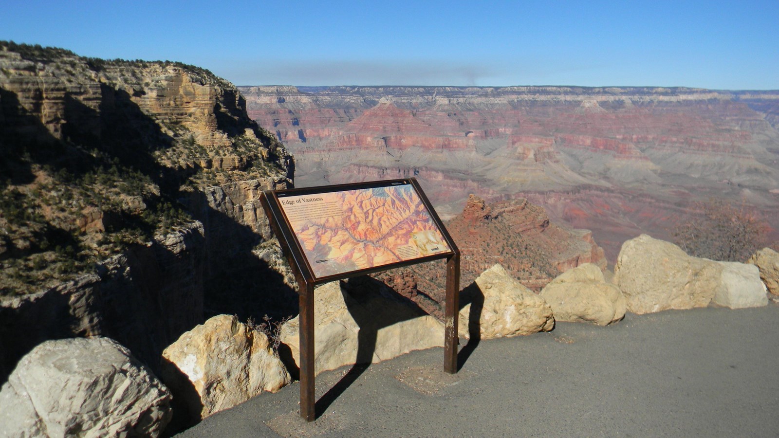 A colorful sign with an aerial view of Grand Canyon stands in front of the canyon itself.