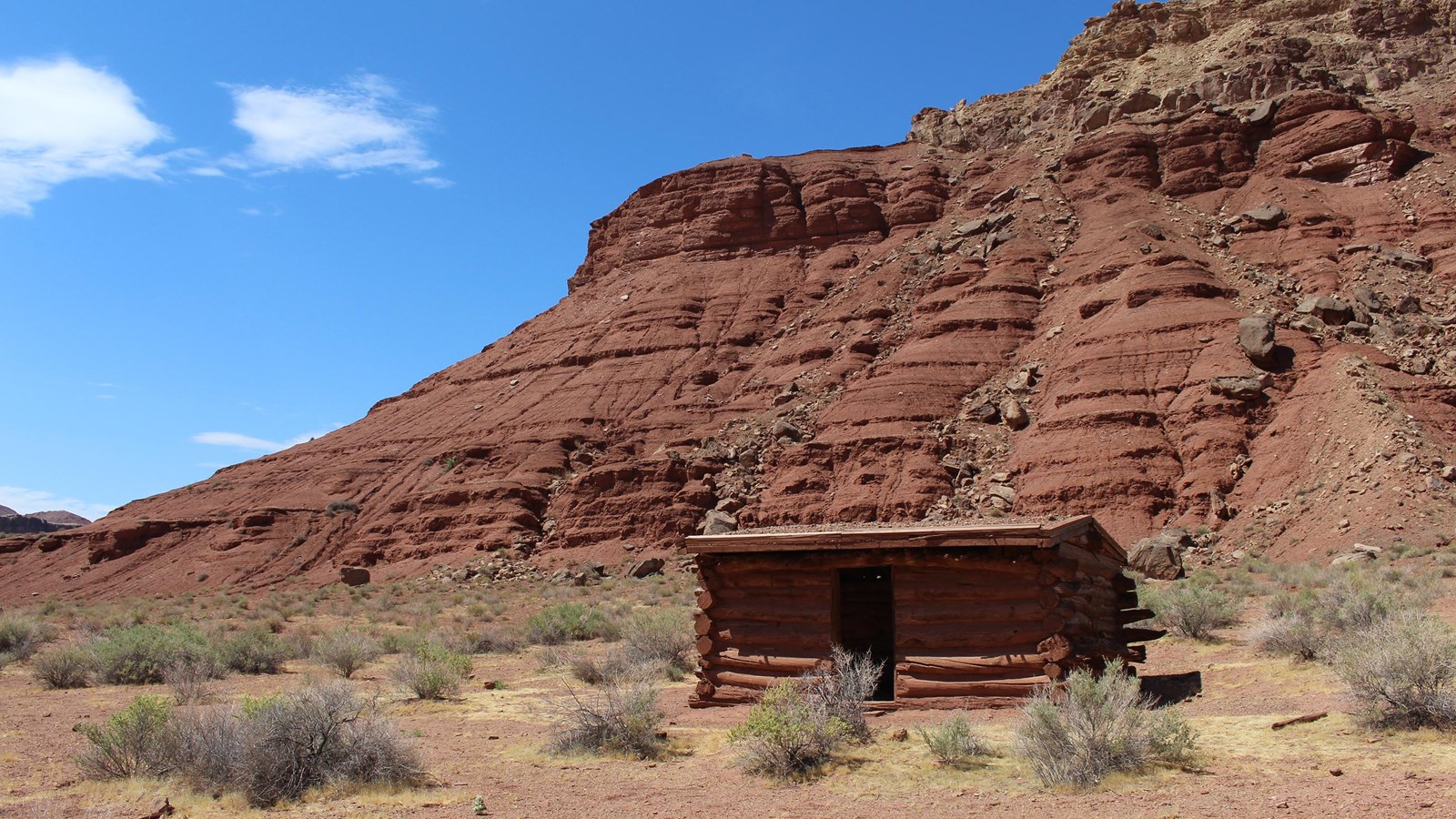 Single story, one room wooden structure surrounded by red desert. Canyons rise in the background.