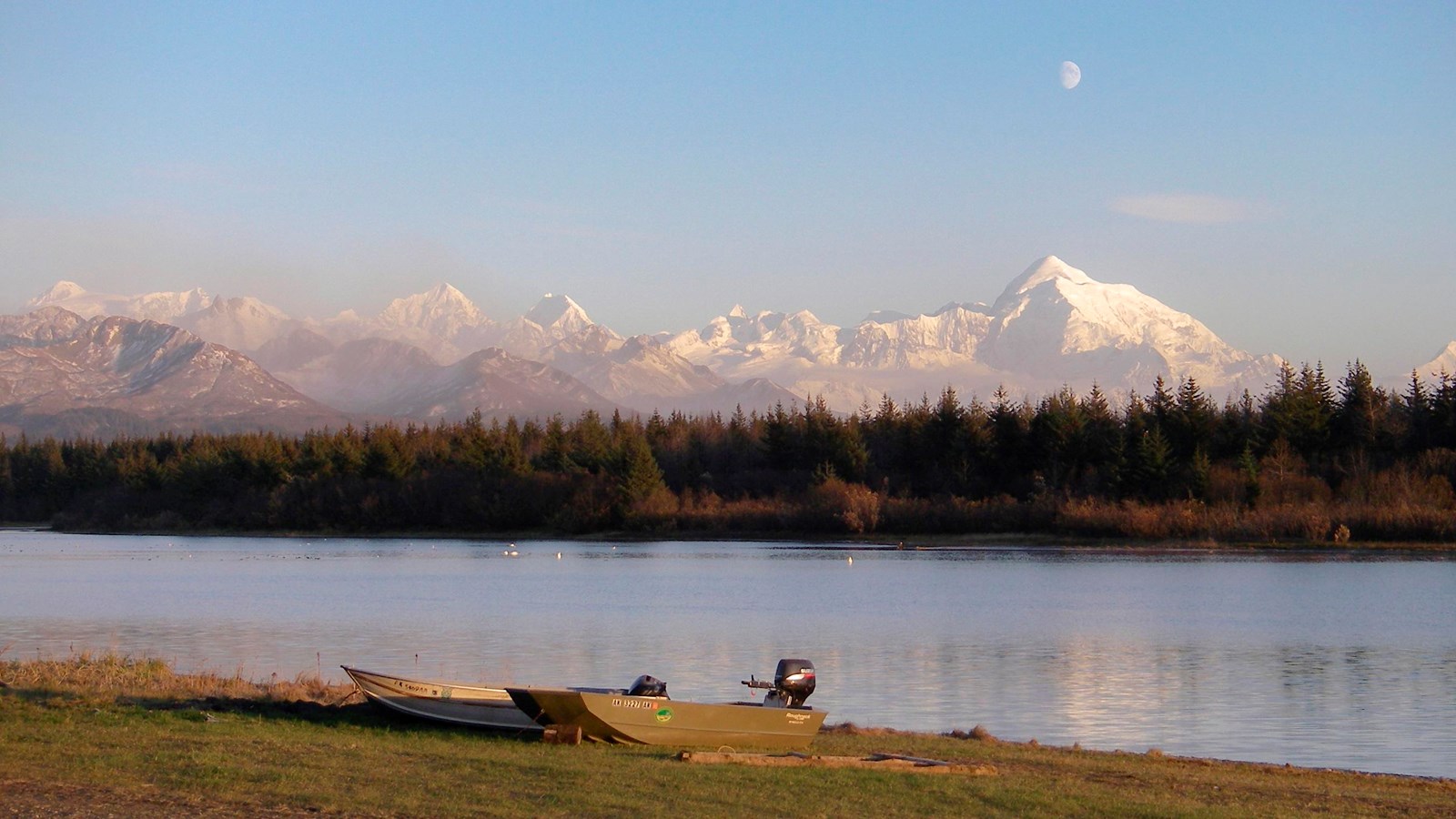 Boats on the shore of a river, distant snowy mountains and a full moon in the background