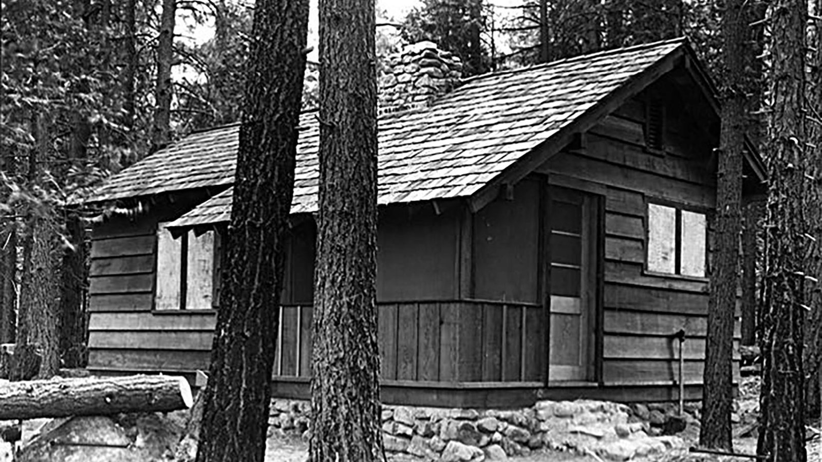 Black and white image of a small, wooden cabin surrounded by trees. 