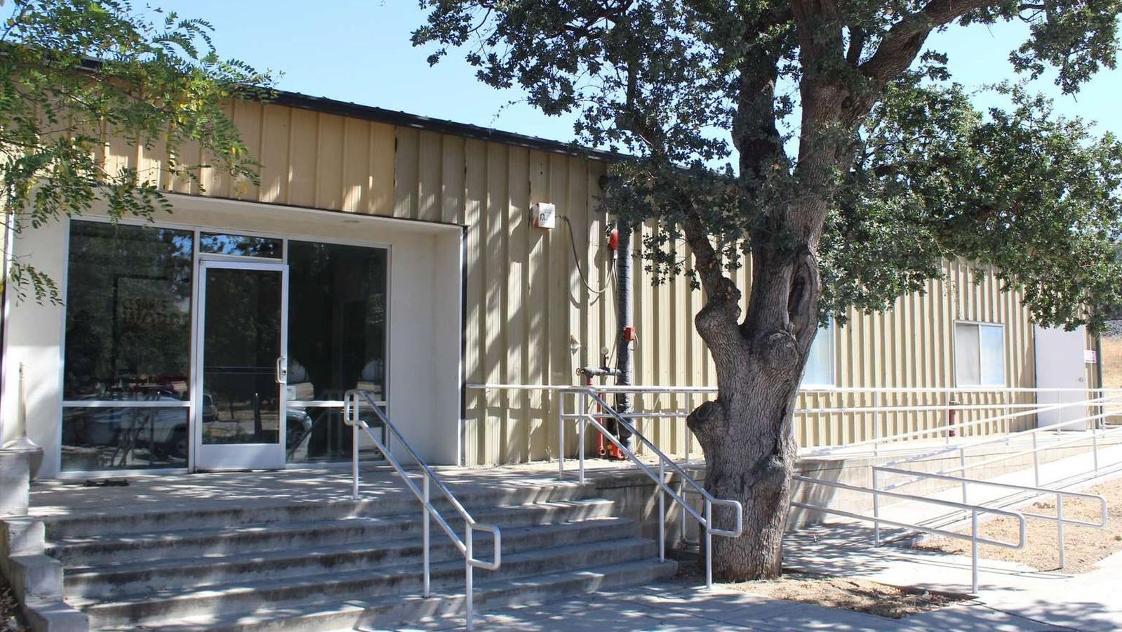 A tan metal rectangular building with stairs and a ramp. There are trees in front of the building.