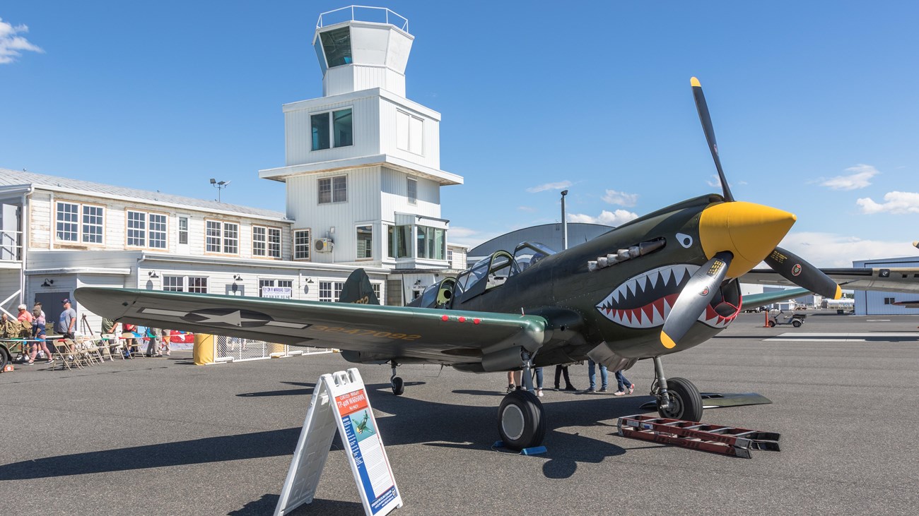 Color photograph of a green airplane with a yellow nose in front of a white building with a square t