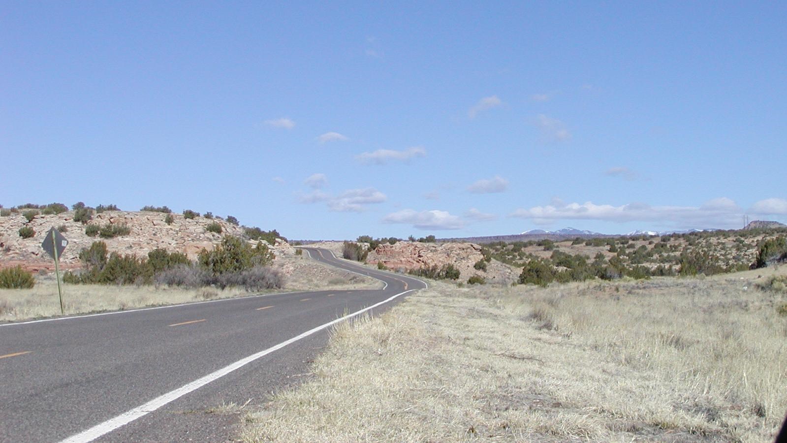 A paved road winds through a field of tan grass. Hills with trees are in the distance.