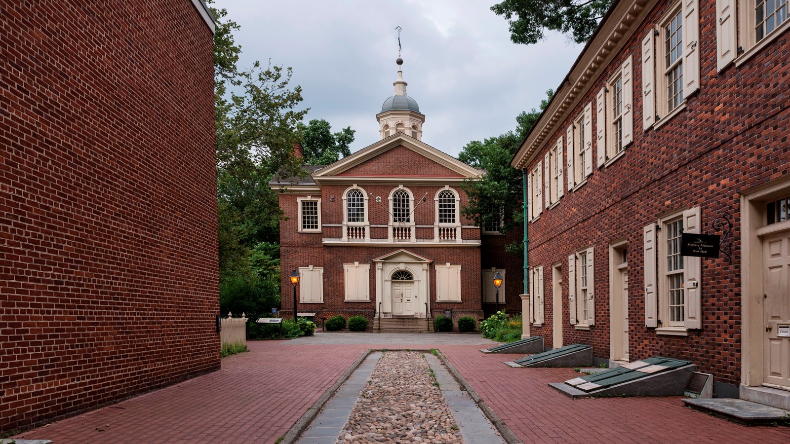 A color photo showing a cobblestone path leading to a two story red brick building with a cupola. .