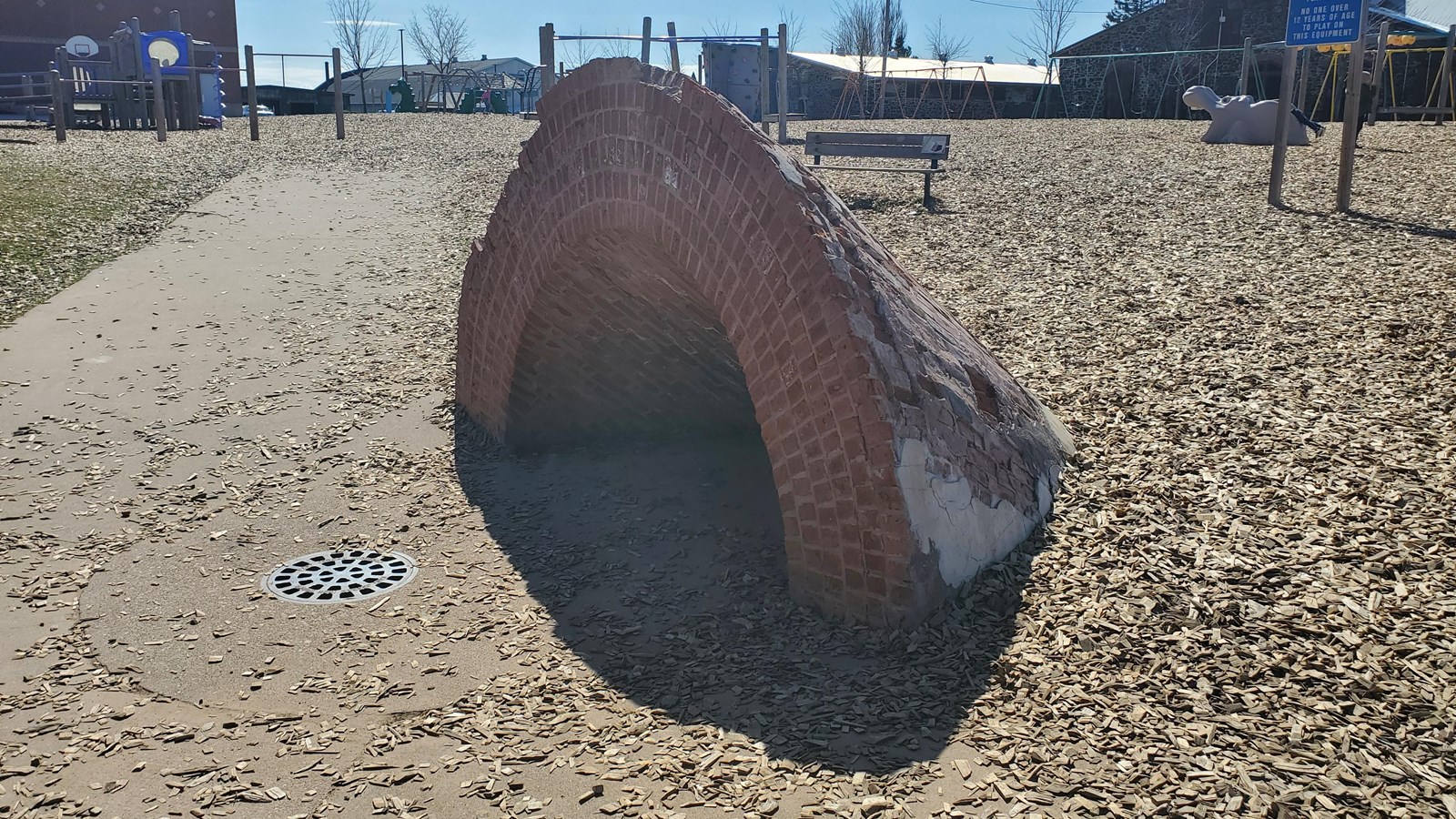 Brick arch rising out of concrete with woodchips and playground equipment in the background. 