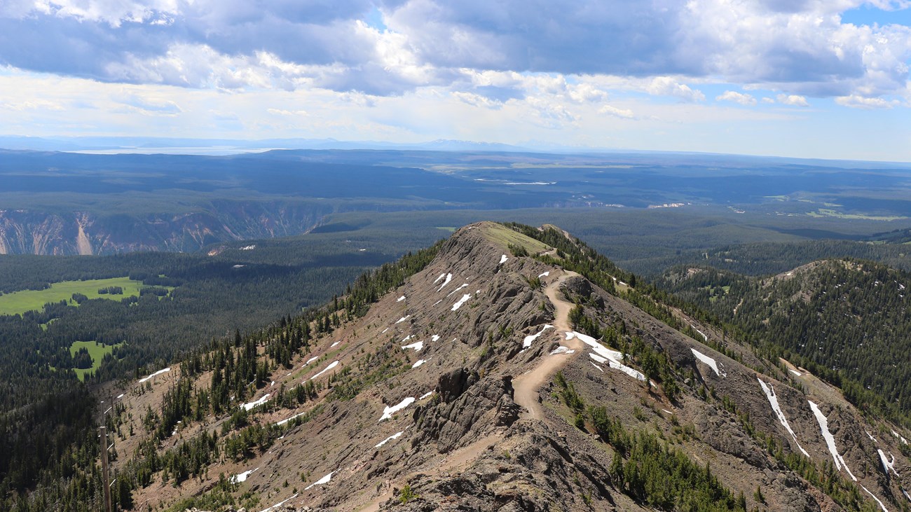 A trail crosses a ridgeline above forests and meadows.