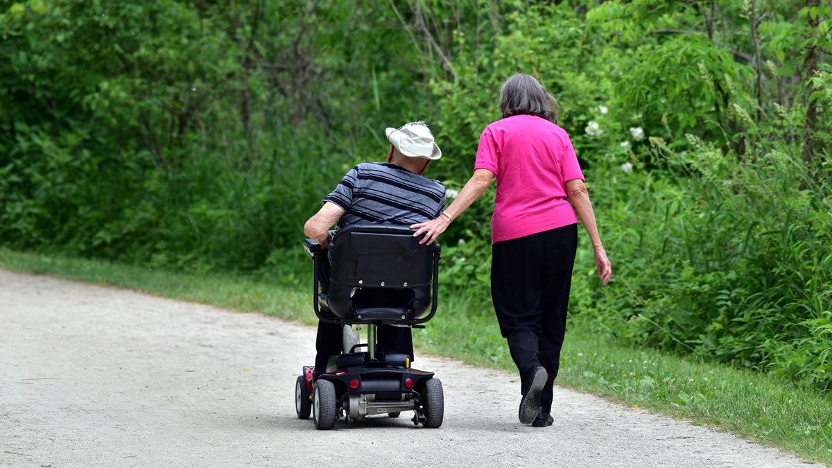 A woman walks beside a man on a flat trail, resting a hand on the back of his motorized wheelchair.