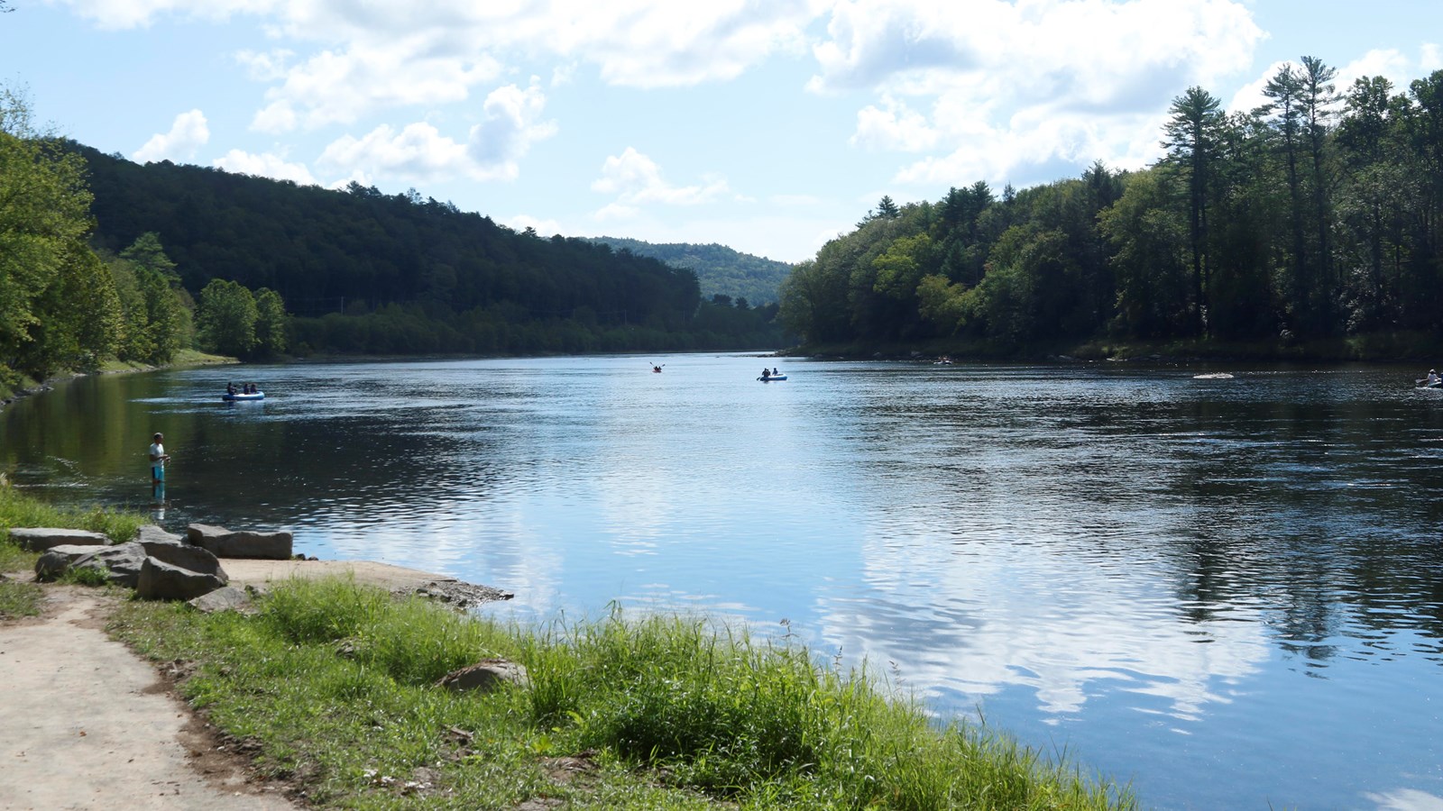 View of paved path leading down to the river. River reflects blue and clouded sky. Hills in distance