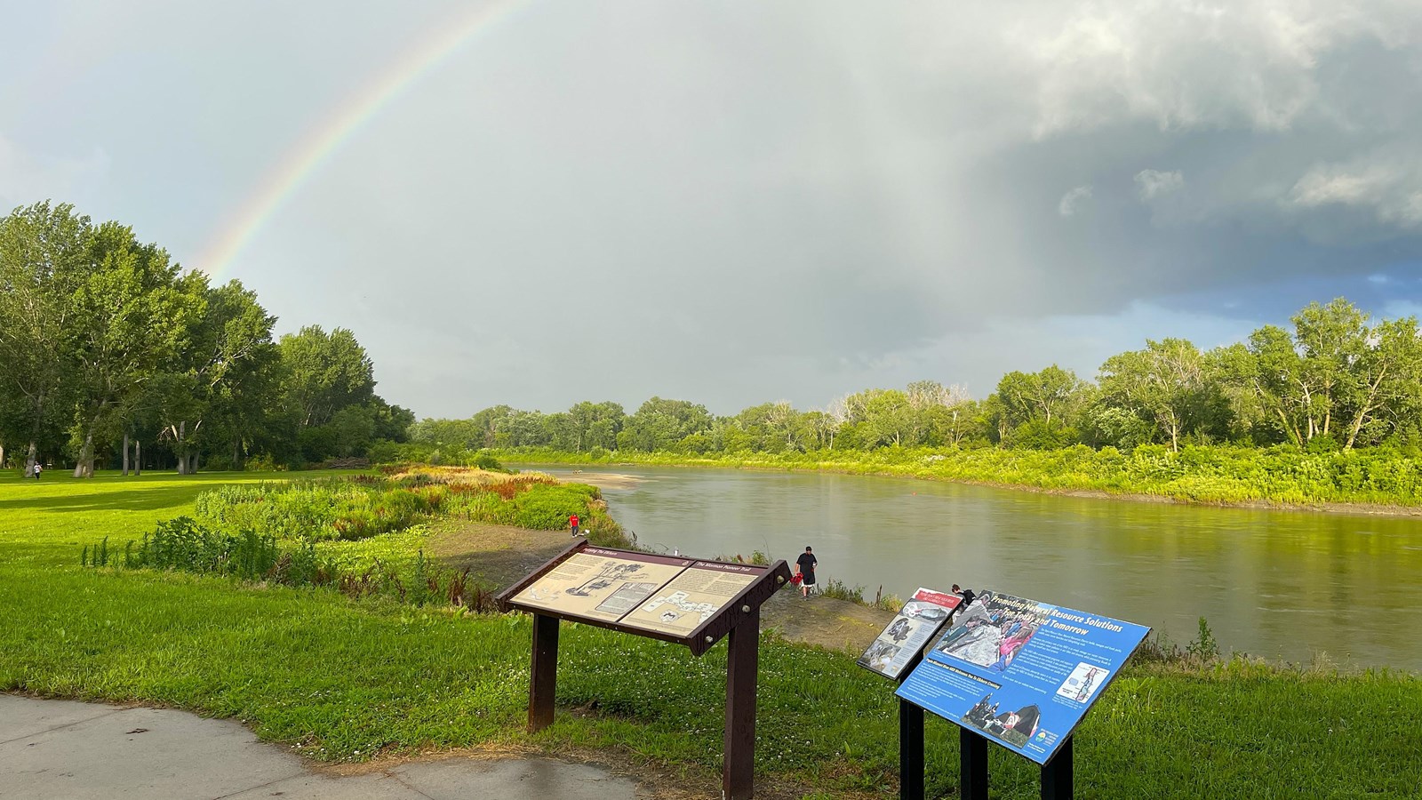 Elkhorn River flows past wayside panels, banks, and wildflowers. A rainbow is towards the left.