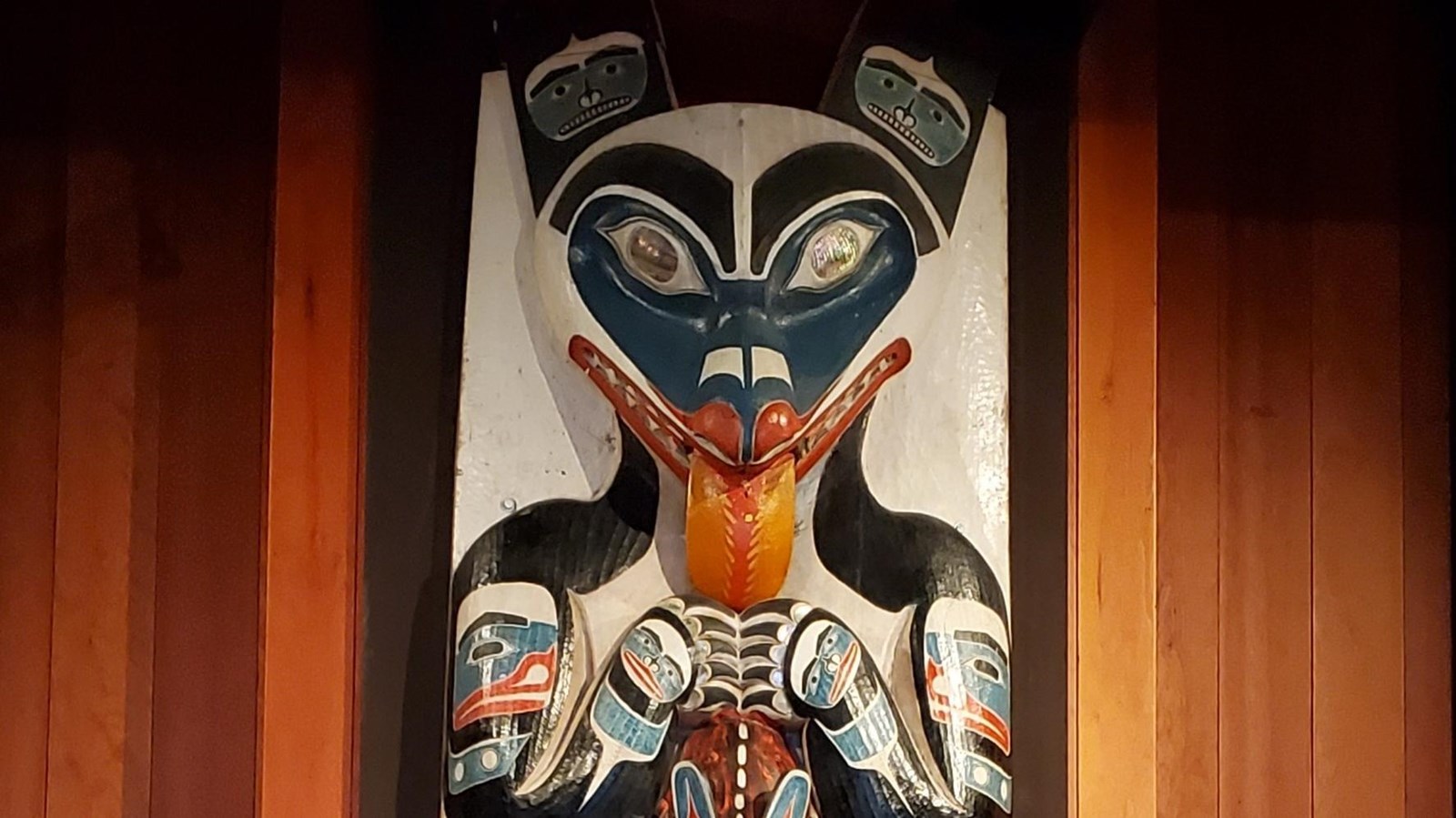 Top portion of a house pole depicting a wolf holding a smaller wolf, its tongue is extended.