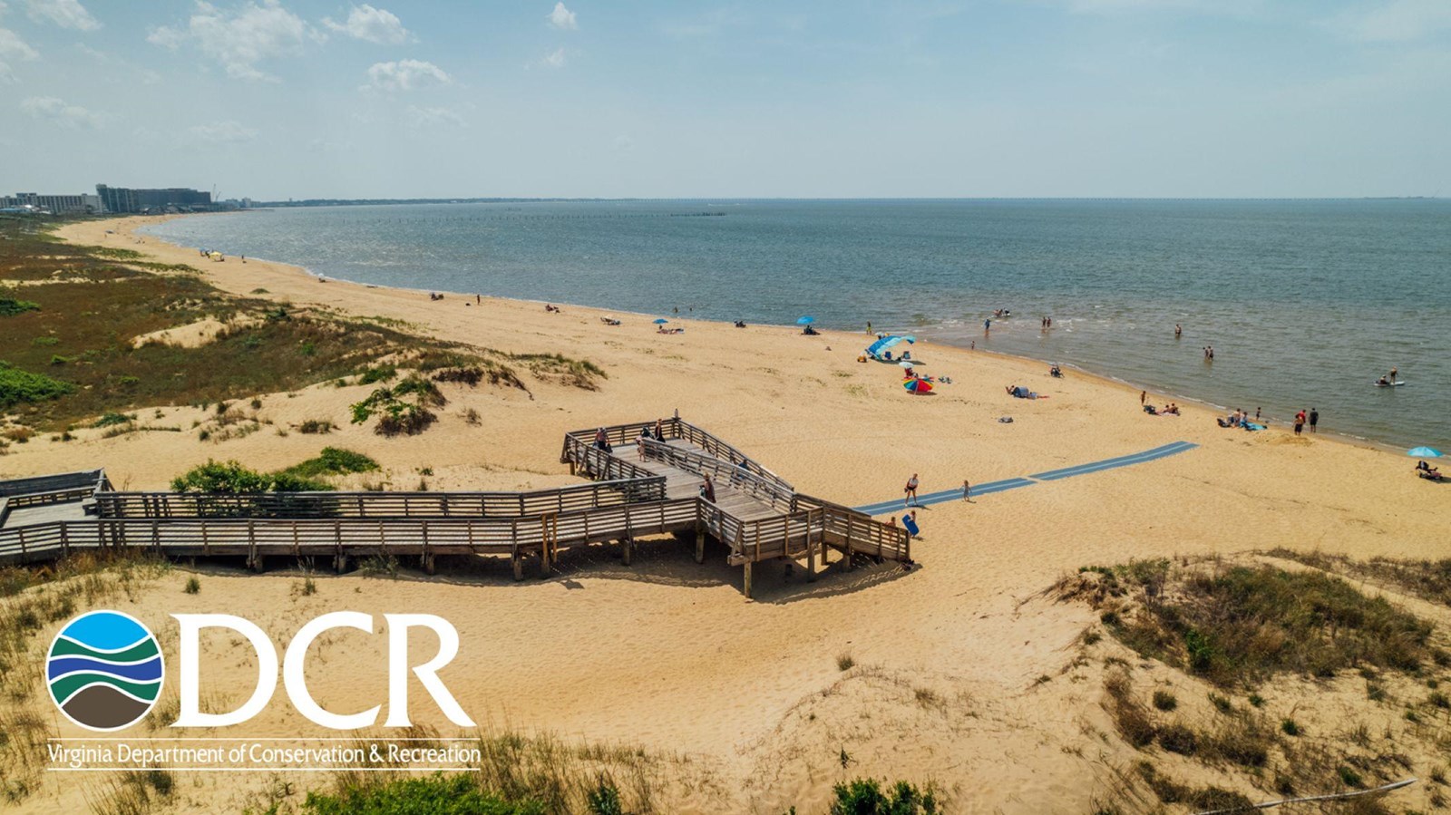 A sandy beach with a wooden boardwalk, colorful umbrellas, and people relaxing under a blue sky.