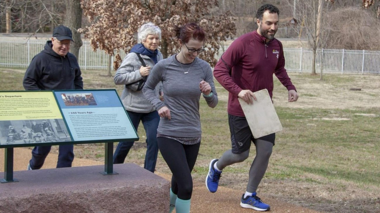 Four people in running gear lightly jogging on concrete with grass in the background.