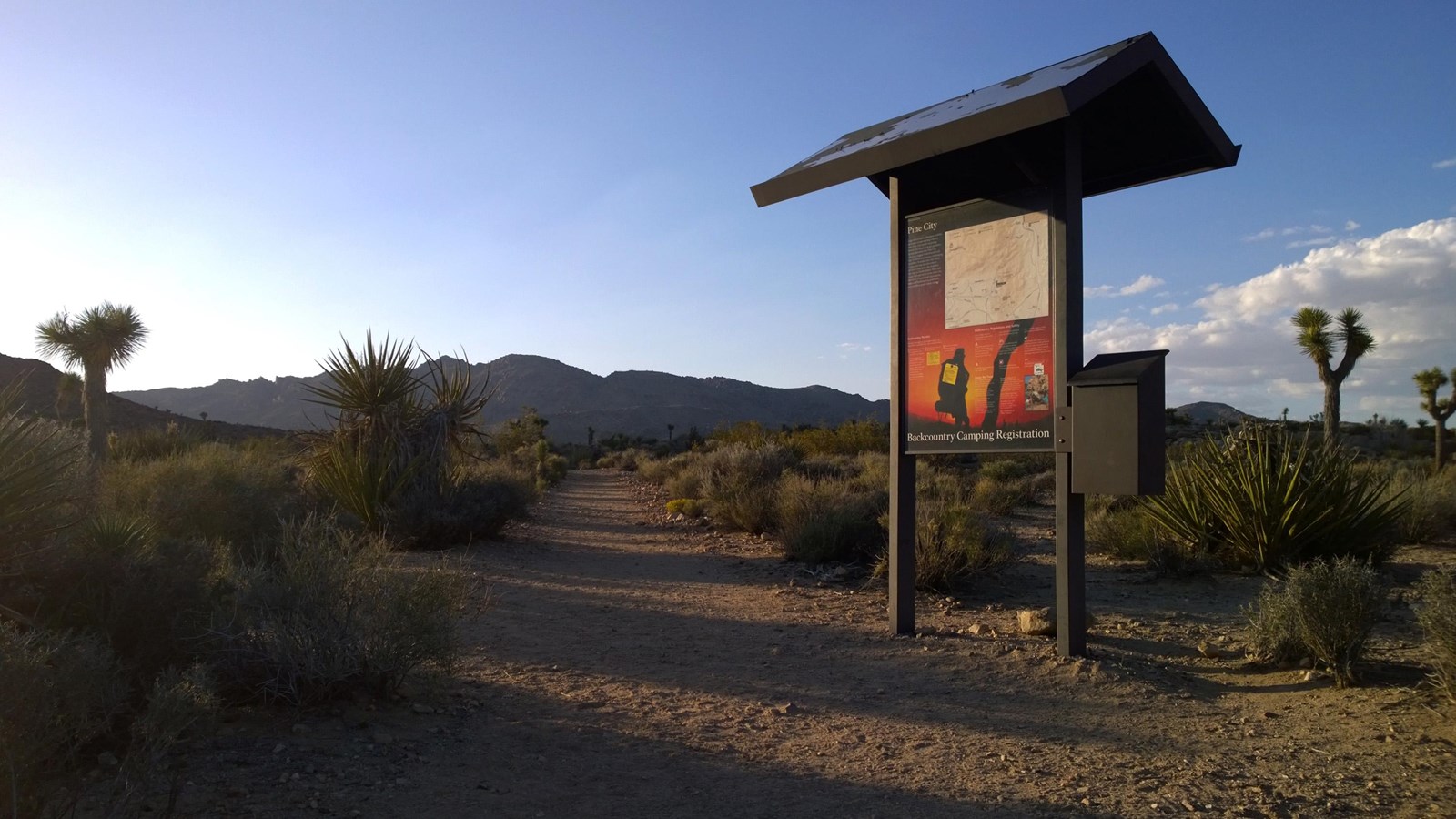 A dirt trail going between desert shrubs past a backpacking bulletin boards towards mountains.
