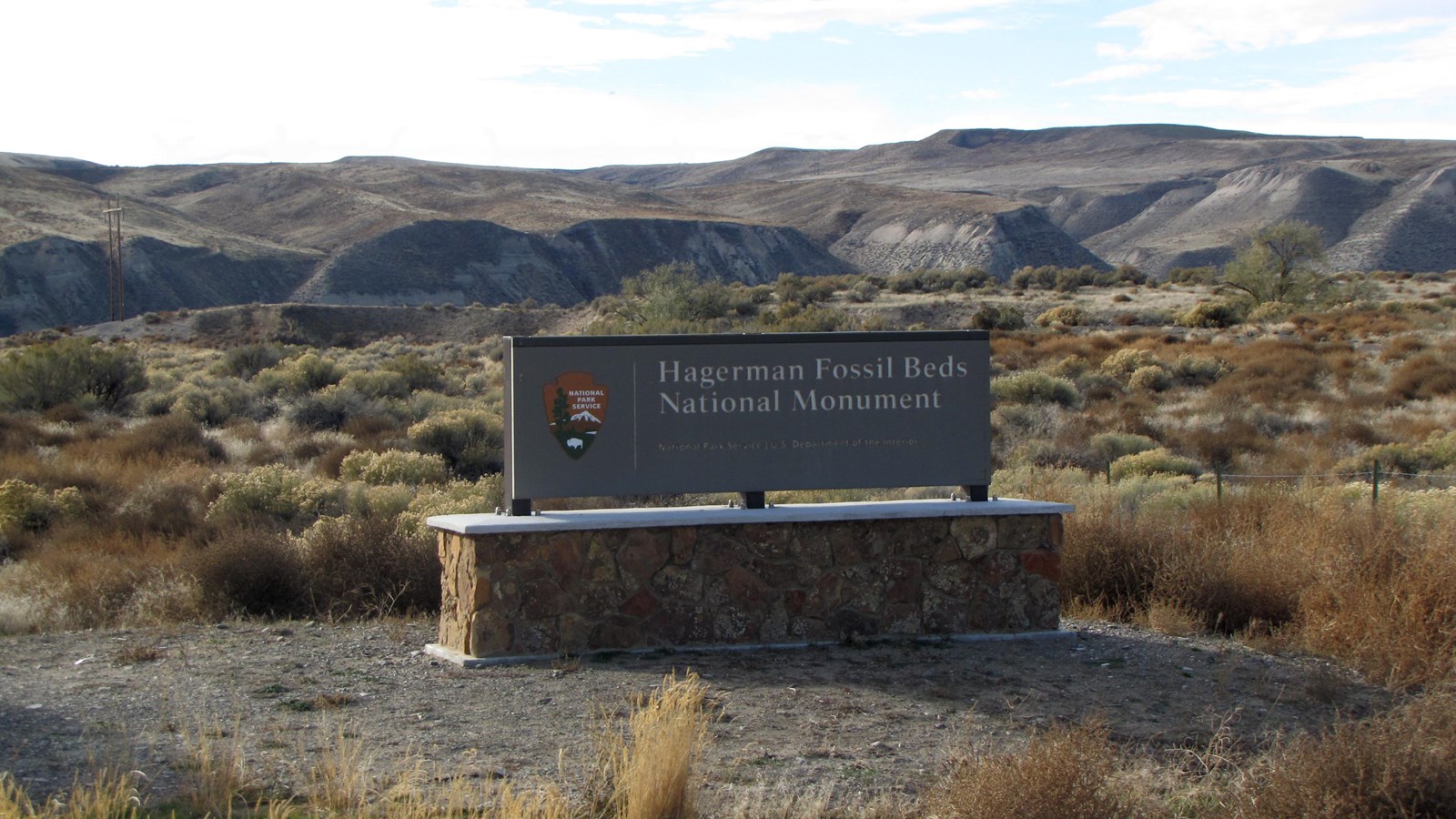 A brown sign introducing the park entrance with large eroded brown hills behind it.