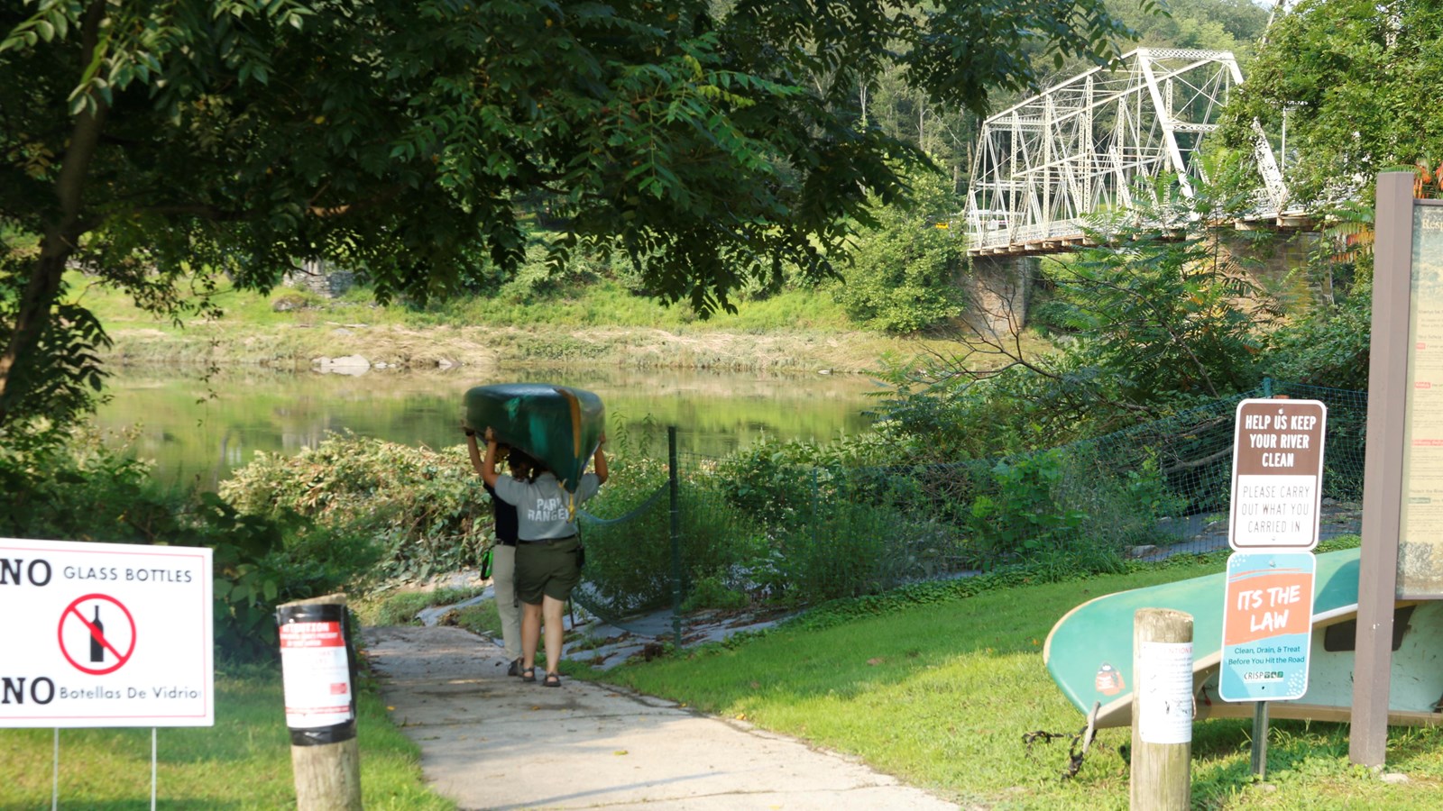 Park rangers hold canoe over heads as walk down paved path towards water. In distance is a bridge.