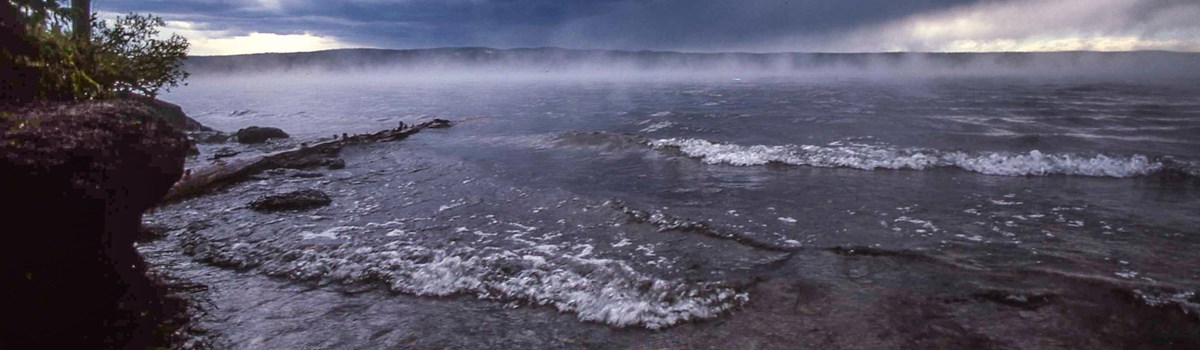 Water crashes on the rocky shore while mist hangs over the lake.