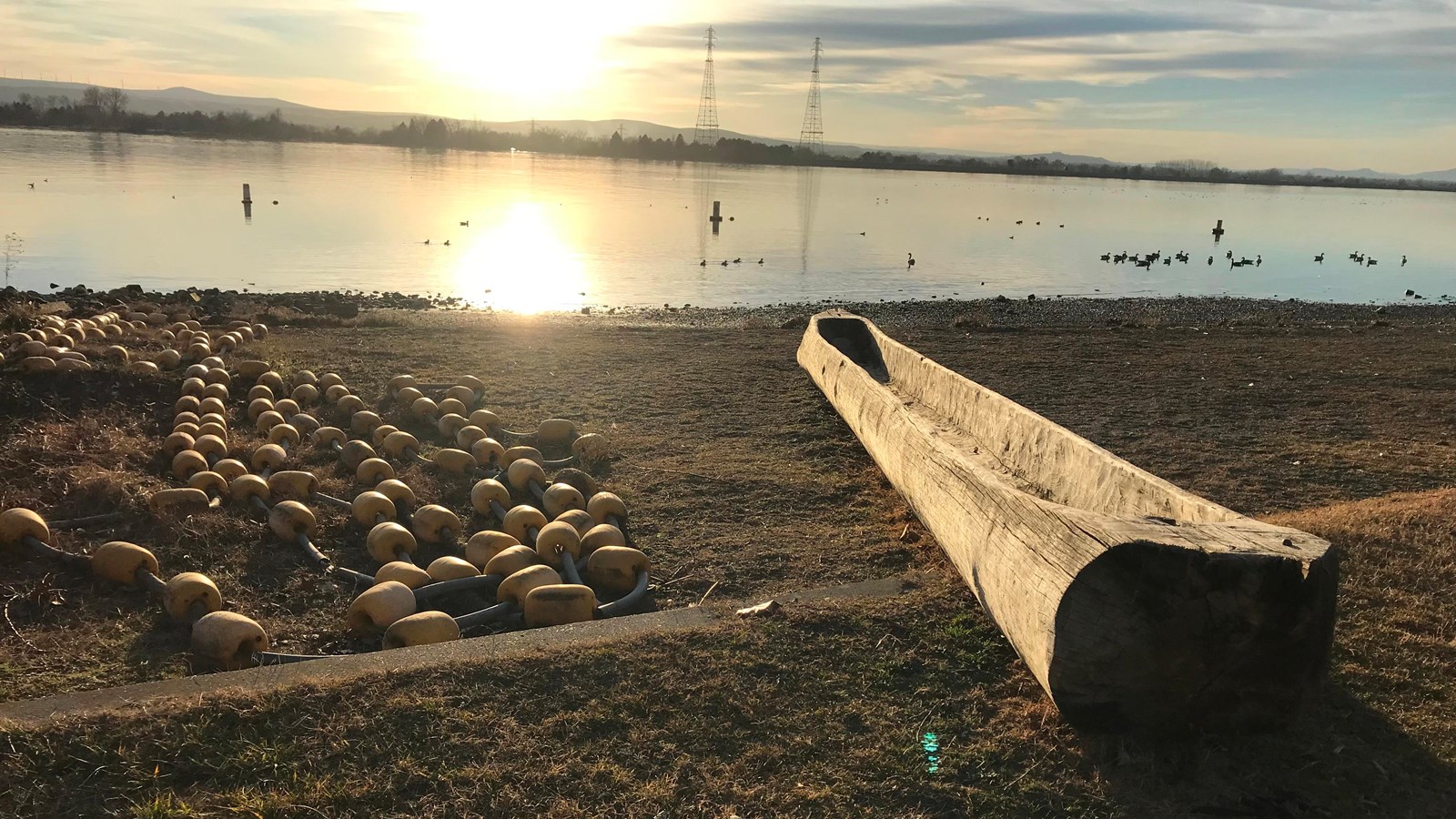 A dugout canoe rests on the bank of a large river at sunset 