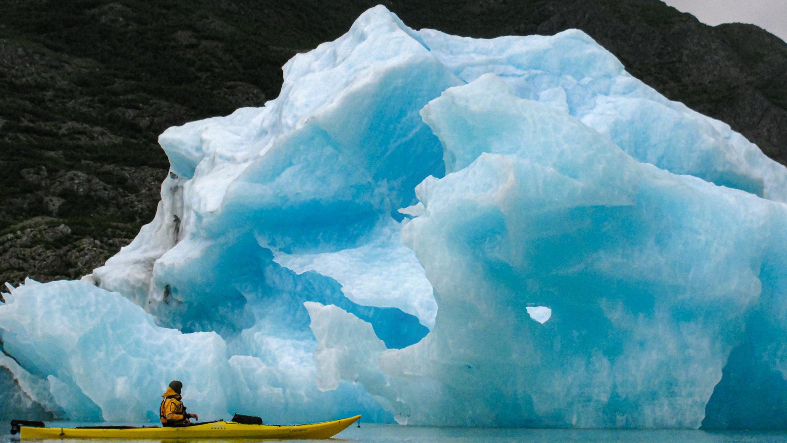 A kayaker paddles by a huge blue iceberg.