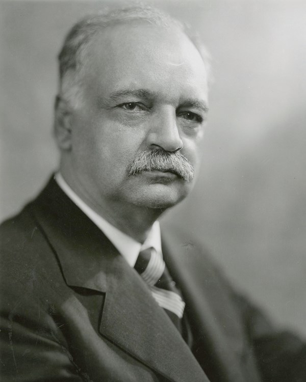 A 1928 bust portrait photo shows a serious gray haired and mustached man in a suit. 