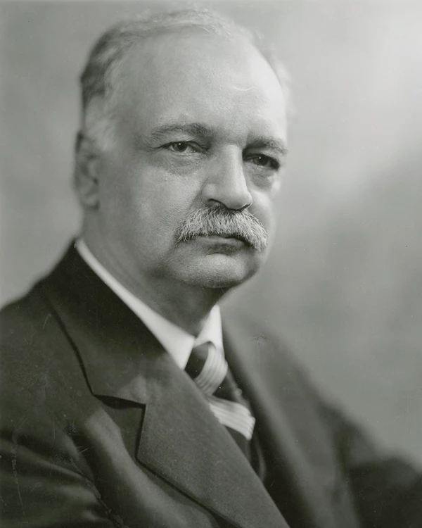 A 1928 bust portrait photo shows a serious gray haired and mustached man in a suit.