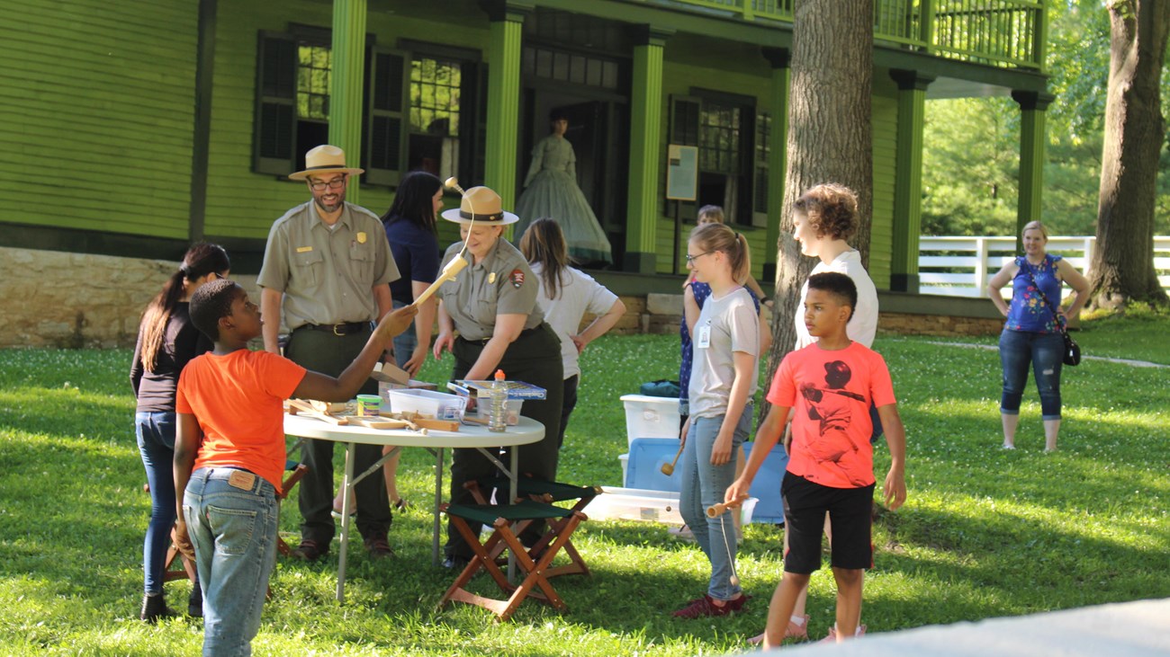 Two park rangers at a table standing alongside five students with a green house in the background.