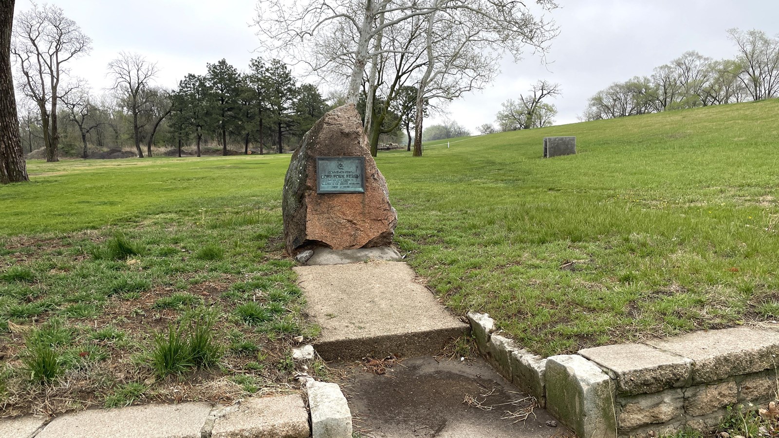 Red granite monument with plaque, surrounded by landscaping blocks, grass, trees, and sidewalk.