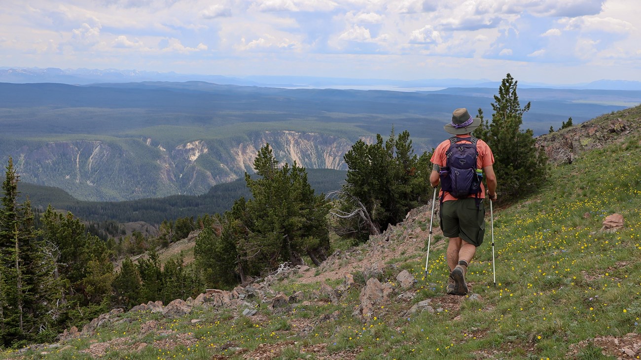 A hiker walks on a mountain slope with a canyon and lake seen in the distance.