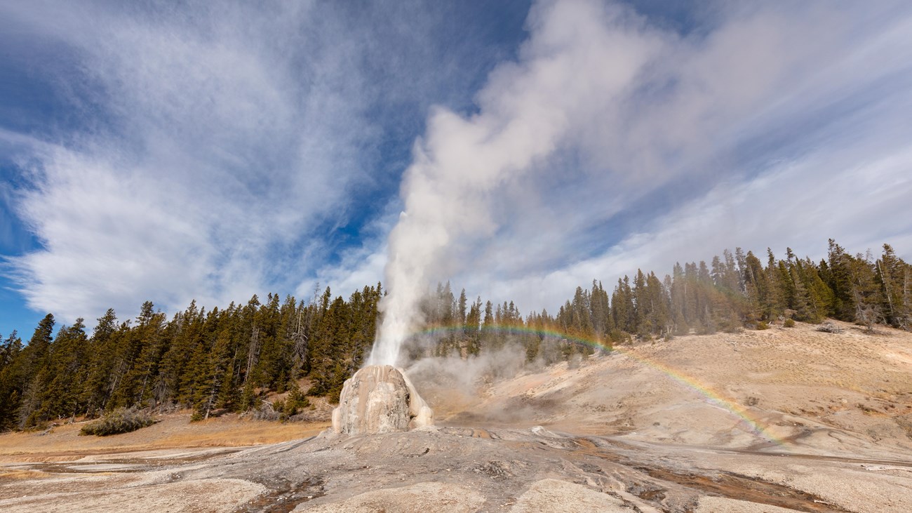 Steam and water erupt from the tan cone of Lone Star Geyser. A rainbow appears in the mist.