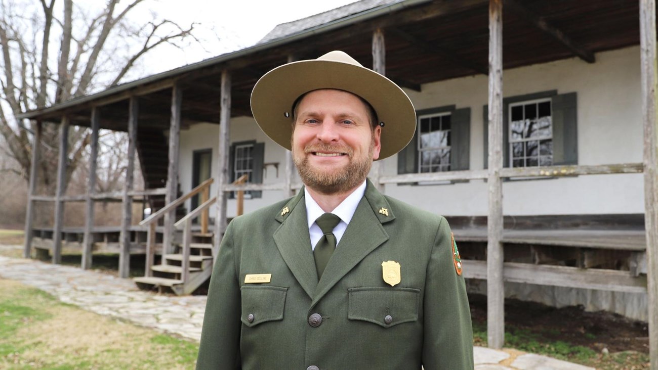 A smiling park ranger stands in front of a historic building