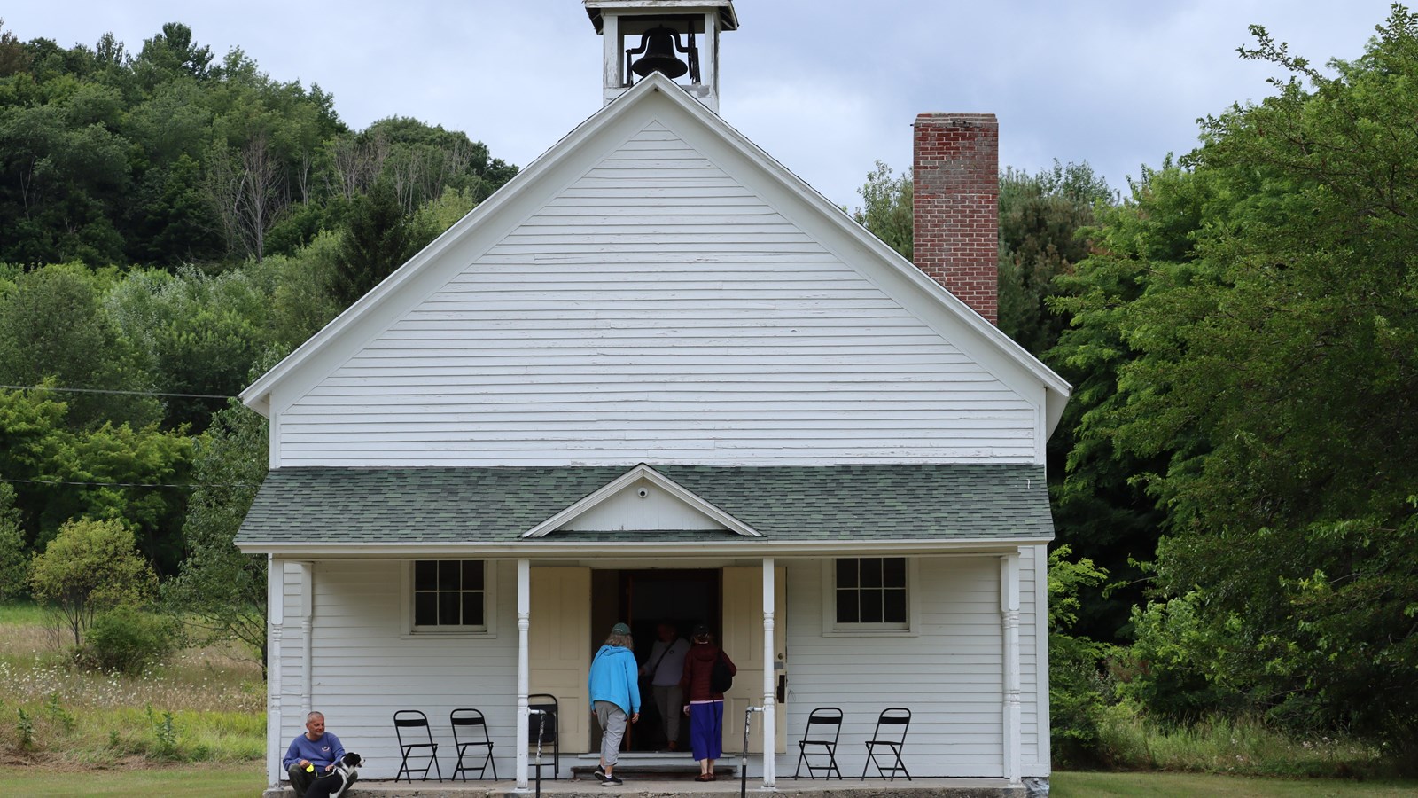 Tall white one-room schoolhouse with a bell tower and open doors. Several adults walking in.