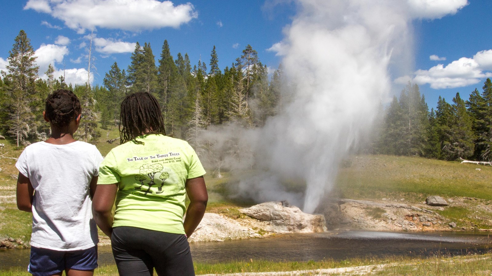 Two young girls stand across the river from an erupting geyser.