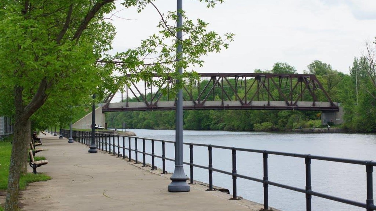 Along a sidewalk, you can see a metal railing and just beyond that, a river and a metal bridge. 